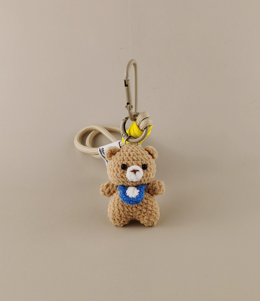 Simple cute bear keychain, with a blue collar and white belly patch in the style of Amigurumi. The background is beige. A yellow banana hangs on the hook of an anime bag or schoolbag, cartoon character design, high resolution, high detail, high quality.