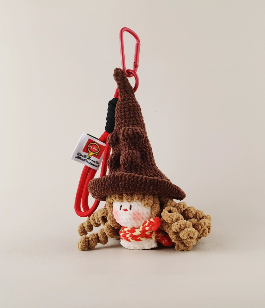  A cute amigurumi brown gnome wearing a witch hat, a red cardigan, and a white tag attached to the back of his head with the text 'ichigo' in Japanese; it is hanging from a clip on a solid background