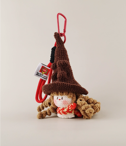  A cute amigurumi brown gnome wearing a witch hat, a red cardigan, and a white tag attached to the back of his head with the text 'ichigo' in Japanese; it is hanging from a clip on a solid background